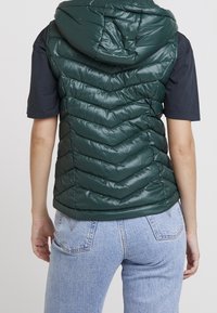 Osoba ubrana w ciemnozieloną pikowaną kamizelkę bez rękawów z kapturem, noszącą czarną koszulę z krótkim rękawem i jasnoniebieskie dżinsy, widziana z tyłu.