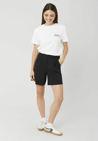 T-shirt en coton blanc avec l'inscription "Sweet or Sour" sur la poitrine, associée à un short noir taille haute. Le mannequin porte des baskets blanches.