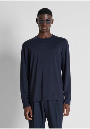 Giovane uomo che indossa occhiali da sole scuri, una camicia a maniche lunghe blu navy e pantaloni blu navy abbinati, in piedi su uno sfondo chiaro e uniforme.