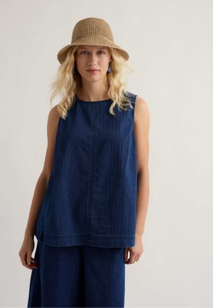 Kobieta o falowanych blond włosach nosi beżowy pleciony kapelusz, bezrękawnik z ciemnoniebieskiego denimu, pasujące szerokie spodnie oraz ciemnoniebieskie kolczyki.