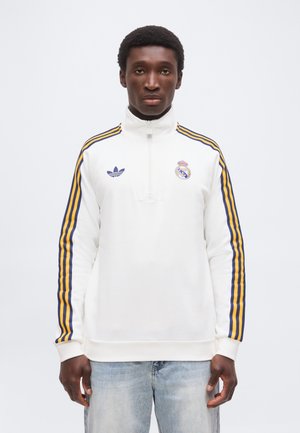 Junger Mann, der ein weißes Real Madrid Viertel-Reißverschluss-Sweatshirt mit blau-gelb gestreiften Ärmeln und hellblauen Jeans trägt, nach vorne gerichtet.