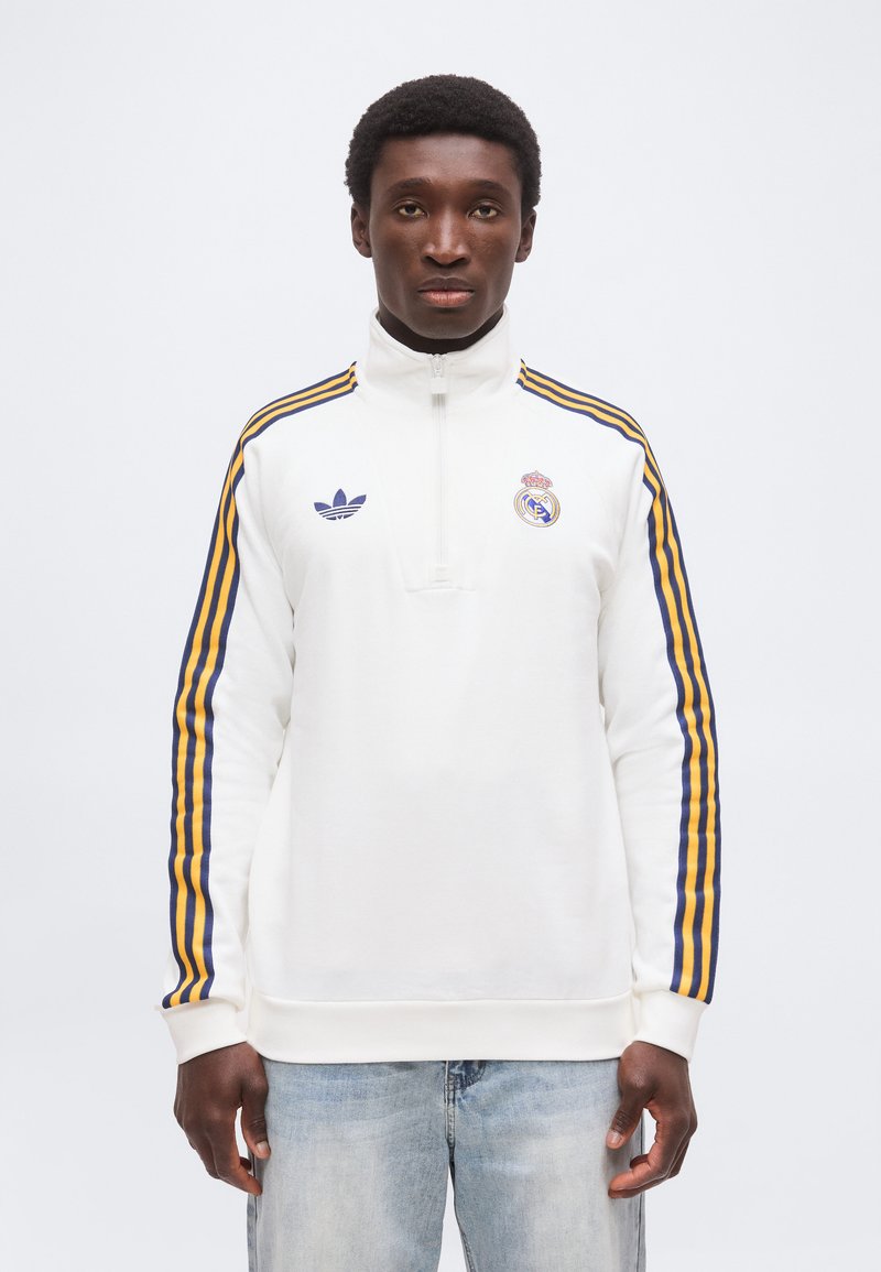 Jeune homme portant un sweat-shirt blanc Real Madrid à fermeture quart-zip avec des manches rayées bleues et jaunes et un jean bleu clair, face à l'avant.