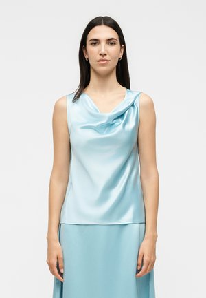 Femme aux longs cheveux foncés portant un top en satin bleu clair sans manches avec un décolleté drapé et une jupe assortie, debout devant un fond uni.