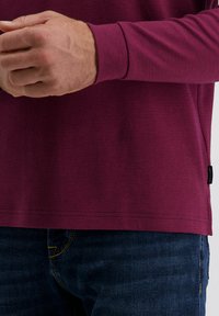 Langärmliges Hemd aus tiefburgunderfarbigem Stoff mit strukturiertem Muster. Der Saum weist Steppdetails auf; kombiniert mit dunklen Jeans.