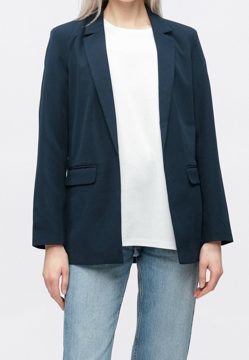 Personne portant un blazer bleu foncé sur une chemise blanche et un jean bleu clair, debout devant un fond blanc uni.