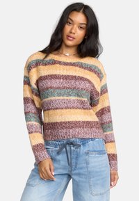 Sweter w paski z dzianiny w odcieniach żółtego, brązowego i teal z luźnym krojem, szerokim dekoltem i prążkowanymi mankietami. W zestawie z jasnoniebieskimi spodniami.