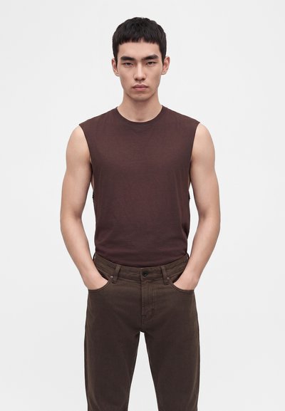 Jeune homme aux cheveux noirs courts portant un débardeur brun foncé et un pantalon marron assorti, les mains dans les poches.