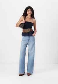 Marinefarget uten ermer topp med sammensatt tekstur, brun bred belte med sirkulært detaljer, lyseblå vide jeans, og brune høye sko.