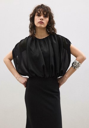 Žena sa kovrčavom smeđom kosom nosi prozirnu crnu bluzu s naborima na rukavima, usku crnu suknju i srebrnu cvjetnu narukvicu, stoji s rukama na bokovima.