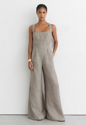 Vrouw met gevlochten haar die een mouwloze, vierkante hals beige linnen jumpsuit met wijde pijpen en een gouden armband draagt, staand tegen een witte muur.
