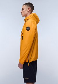 Yellow, waterproof jacket with a hood, elastic cuffs, and front pockets. Features a round logo patch on the left sleeve. Styled with dark shorts.
