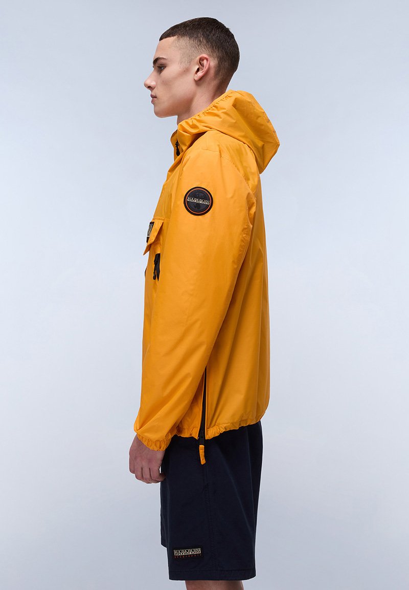 Gelbe, wasserdichte Jacke mit Kapuze, elastischen Bündchen und frontalen Taschen. Verfügt über ein rundes Logo-Patch am linken Ärmel. Kombiniert mit dunklen Shorts.