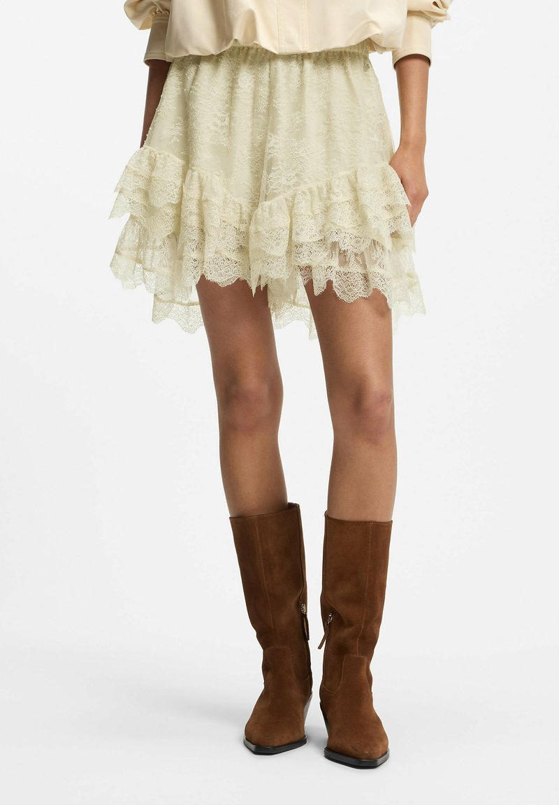 Mujer con mini falda de encaje color crema con volantes combinada con botas de ante marrón hasta la mitad de la pantorrilla, de pie sobre un fondo blanco.