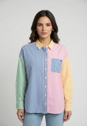 Femme portant une chemise à boutons à rayures verticales multicolores avec des panneaux verts, bleus, roses et jaunes, ainsi qu'un jean bleu clair sur un fond uni.