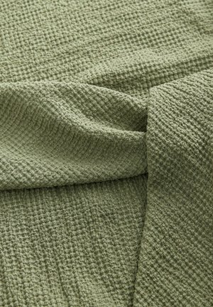 Teksturowany zielony materiał z wzorzystym splotem, charakteryzujący się miękkim, pomarszczonym wyglądem. Materiał opada delikatnie, z widocznymi fałdami.