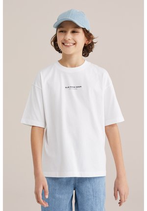 Tânăr zâmbitor purtând o șapcă albastru deschis, un tricou alb cu inscripția "Blue Ridge Denim" și blugi albastru deschis, pe fundal bej.