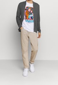 Grå stickad kofta över en vit t-shirt med grafik, beige byxor och vita sneakers. T-shirten har färggård, abstrakt konstverk.