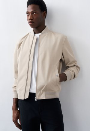 Young man wearing a beige bomber jacket over a white shirt, standing with one hand in pocket, looking to the side against a plain white wall.