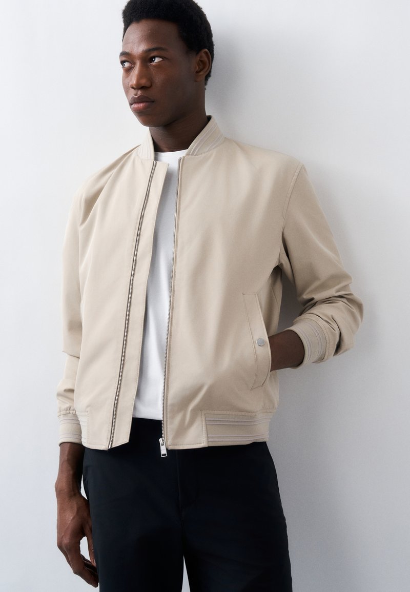 Jeune homme portant une veste bomber beige sur une chemise blanche, debout avec une main dans la poche, regardant sur le côté contre un mur blanc uni.