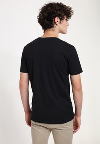 Black cotton t-shirt with a crew neck, short sleeves, and a straight cut, worn with light-colored pants, shown from the back.