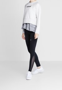 Sweatshirt Nike gris clair avec logo noir, superposé à un haut gris à motif ; associé à un legging noir avec panneaux en mesh. Baskets blanches.