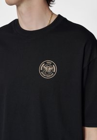 Svart bomulls T-shirt med en rund beige broderad logotyp med en bi, lagerblad och "1923 SPORTSWEAR". Klassisk crew neck design.