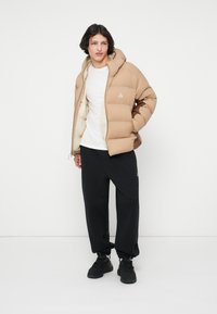 Beige dunjakke med hætte, med lynlås foran og et lille logo på brystet. Sæt sammen med sorte sweatpants og sorte sneakers.