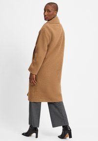 Bruine, geweven wolcoat met een geknipte kraag, zijzakken en een relaxte pasvorm, gecombineerd met grijze, wijde broekspijpen en zwarte enkellaarsjes.
