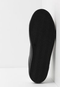 Suola di sneaker nera con un motivo a zigzag, posizionata verticalmente su uno sfondo bianco con angoli.