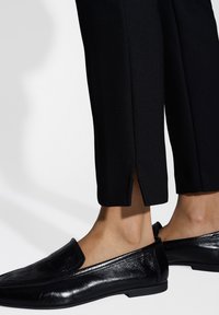 Schwarze maßgeschneiderte Hose mit seitlichem Schlitz und glänzender Verarbeitung, kombiniert mit glänzenden schwarzen Slippern aus glattem Leder.