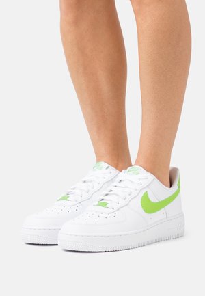 Jambes portant des baskets blanches avec le logo Nike en vert vif et des détails assortis, debout sur un fond gris clair uni.