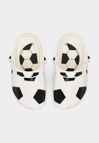 Λευκά clogs χωρίς κορδόνια με μαύρα πεντάγωνα και εξάγωνα μοτίβα που θυμίζουν κλασική μπάλα ποδοσφαίρου, εμφανιζόμενα από πάνω σε απλό φόντο.