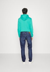 Felpa turchese con tasca a marsupio frontale, abbinata a jeans in denim blu scuro e sneakers bianche, vista da dietro.
