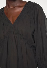 Personne à la peau foncée portant un chemisier noir ample avec un décolleté en V et des manches longues bouffantes resserrées aux poignets.