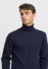 Giovane uomo con capelli castani corti che indossa un dolcevita blu navy, guarda verso il basso e a sinistra su uno sfondo semplice.