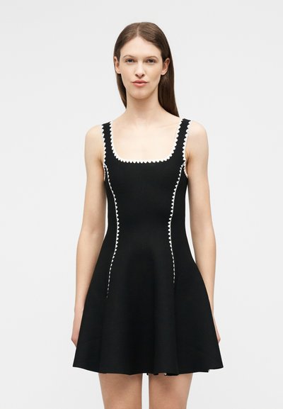 Femme portant une robe noire sans manches avec un bord festonné blanc le long de l'encolure, des emmanchures et des coutures verticales, debout devant un fond uni.