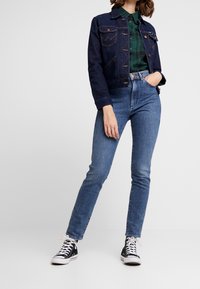 Giubbotto di denim, blu scuro, con tasche sul petto e dettagli di cucitura. Camicia a quadri verdi sotto, abbinata a jeans blu chiaro e scarpe da ginnastica nere.