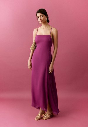 Vrouw staand tegen een roze achtergrond, gekleed in een mouweloze magentakleurige maxi-jurk, gouden oorbellen, sandalen met open tenen, en ze houdt een gouden tas met franjes vast.