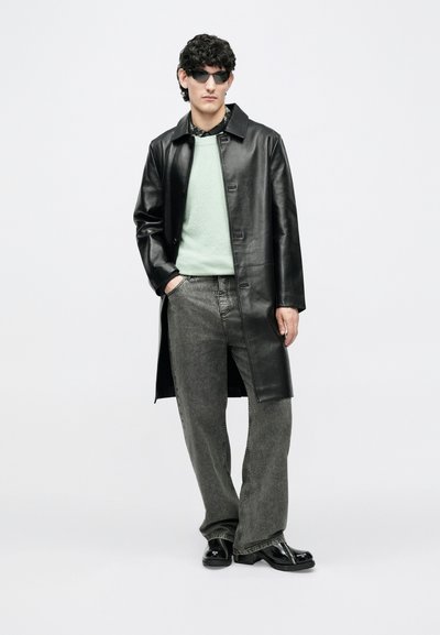 Mees, kes kannab mustast nahast mantlit, mündirohelist sviitrit, halli laia säärega teksapükse, musti saapaid ja päikeseprille, seisab valge tausta ees.