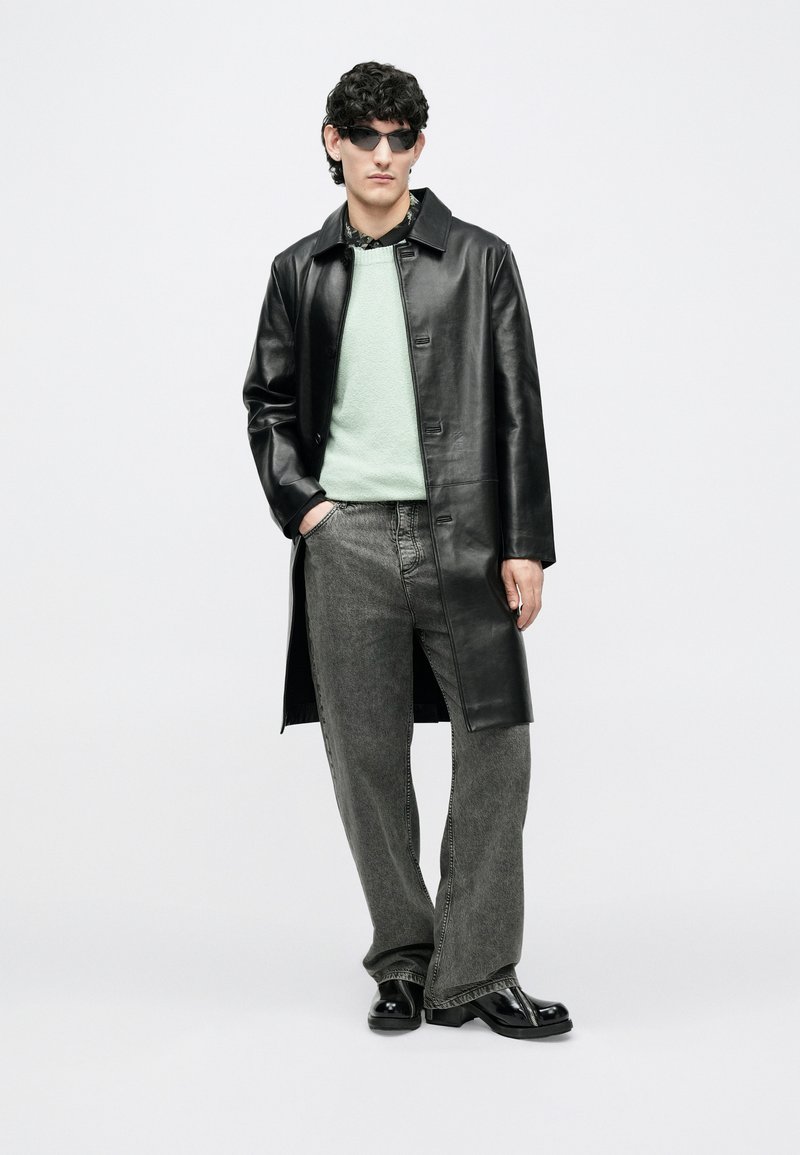 Homme portant un manteau en cuir noir, un pull menthe, un jean gris à jambes larges, des bottes noires et des lunettes de soleil, debout devant un fond blanc.