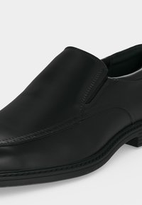 Μαύρο δερμάτινο παπούτσι τύπου slip-on με στρογγυλεμένη μύτη, χαμηλό τακούνι, ελαστικές πλευρικές επιφάνειες και διακοσμητικές ραφές στην λεία επιφάνεια.