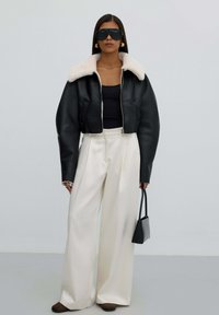 Zwart leren cropped jack met witte nepbont kraag, gecombineerd met wijde witte broek en een kleine zwarte handtas. Model draagt oversized zonnebril.