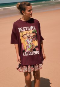 Camiseta oversized burdeos con estampado gráfico que muestra a un hombre con sombrero sombrero. Combinada con una falda con volantes y estampado floral. Fondo de playa con arena.