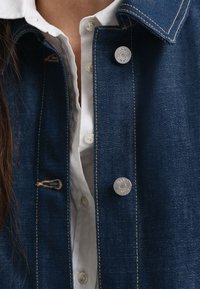 Dongerijakke med metallknapper, mørkeblå stoff, synlige sømmer, og en hvit skjorte under. Teksturet finish og designdetaljer.