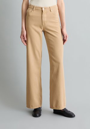 Frau trägt beige weit geschnittene Hose und schwarze Loafer, Hände entspannt an den Seiten, vor schlichtem grauem Hintergrund.