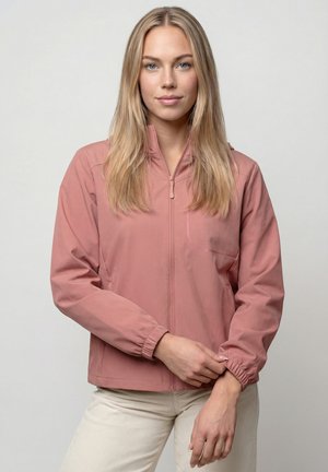 Junge Frau, die den Ärmel einer staubig rosa Reißverschlussjacke hochzieht, trägt hellbeige Hose, steht vor einem schlichten hellen Hintergrund.