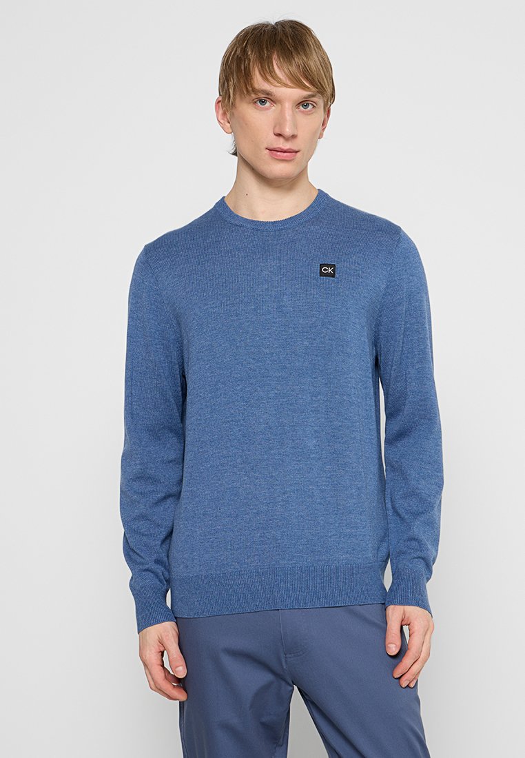 Pull en tricot bleu à manches longues. Encolure ronde. Présente un petit logo CK noir sur la poitrine. Texture douce, poignets et ourlet côtelés.