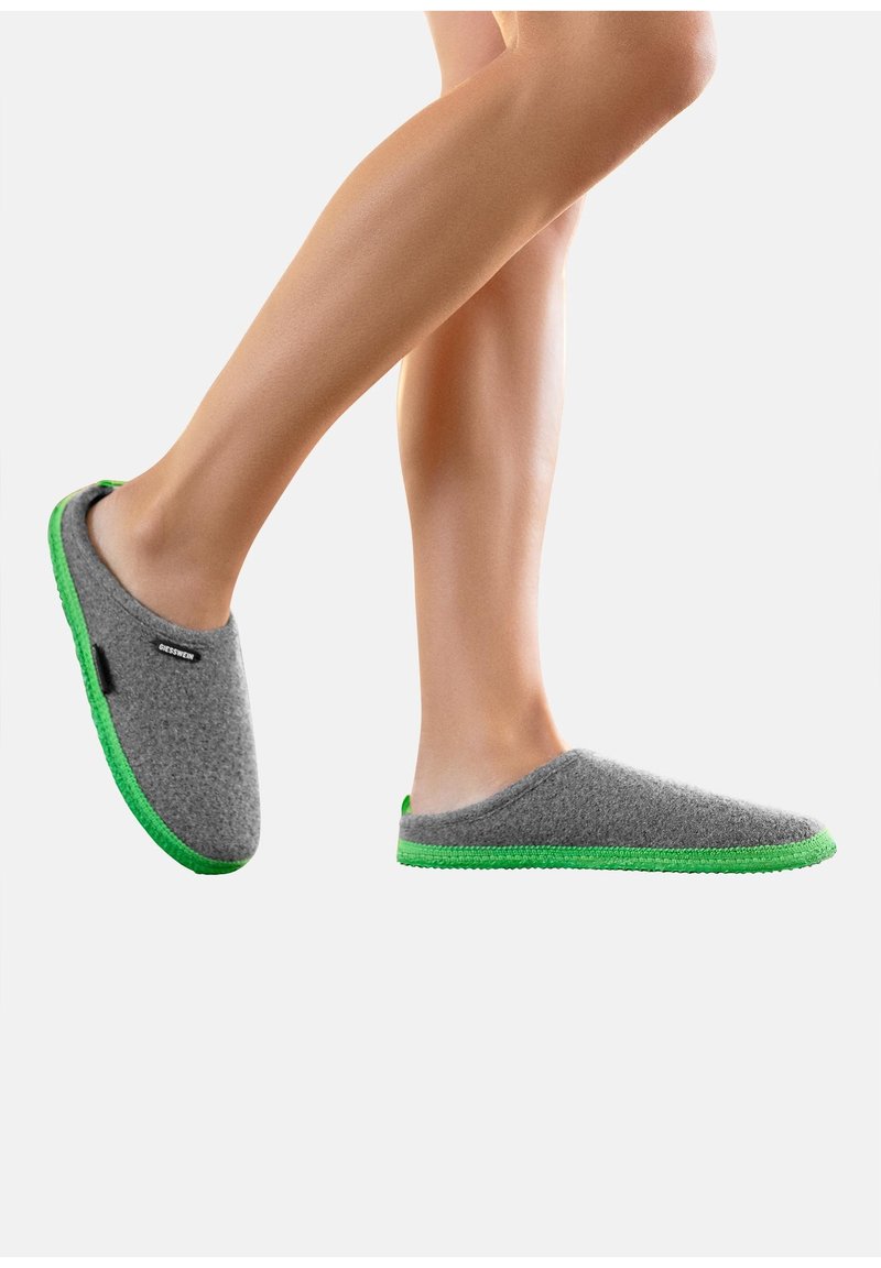 Pieds chaussés de chaussons en feutre gris avec des semelles vert vif sur un fond blanc, un pied légèrement levé comme en train de marcher.