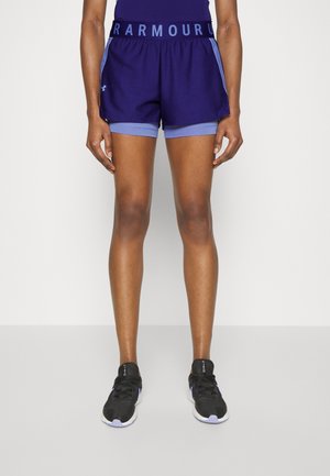 Person iført mørkeblå lagdelte atletikshorts og sorte løbesko, stående mod en ensfarvet lys baggrund.