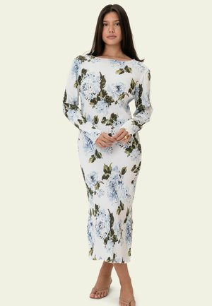 Kvinde står med hænderne foldet, iført en hvid midi kjole med blåt og grønt blomsterprint og beige åbne sandaletter, neutral baggrund.